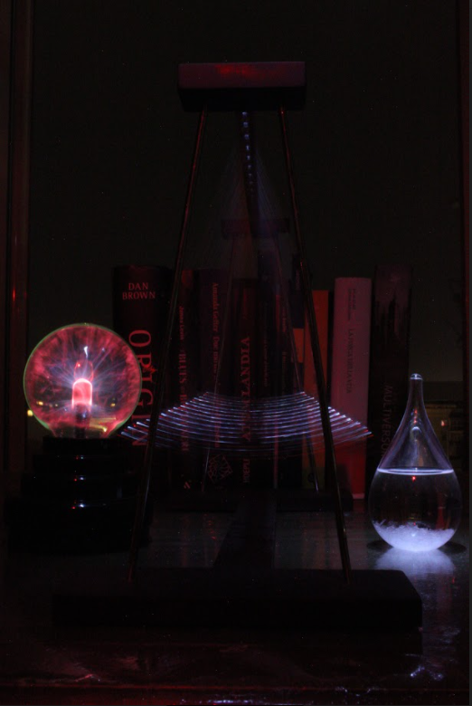 A photograph of a pendulum with a glowing red globe on the left side and a teardrop-shaped flask with water on the right side. In the background is a bookshelf filled with books.